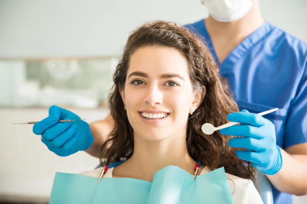 A smiling woman in a dental chair with a dentist or assistant behind her holding tools.