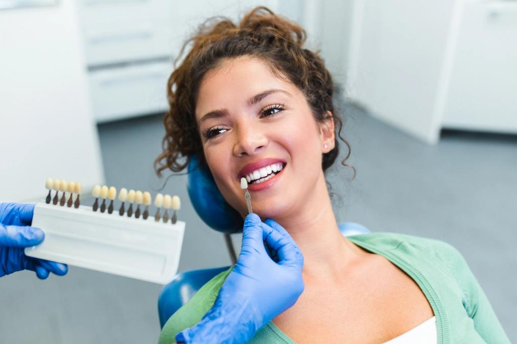 A dental professional compares a smiling woman's tooth shade with a color guide.