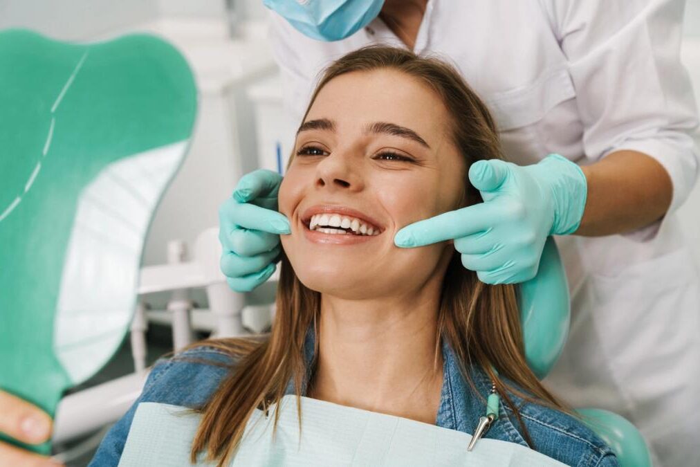 Smiling woman in a dental chair views her bright teeth in a mirror, as a gloved dental professional points to her smile.