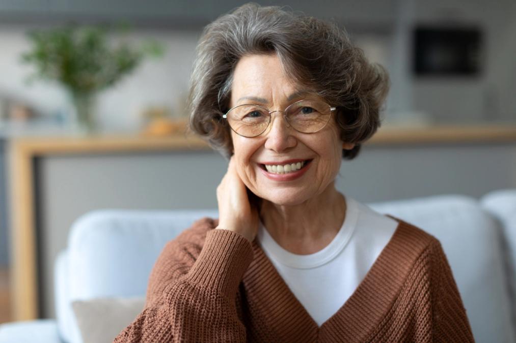 A smiling elderly woman with glasses, hand resting on her neck.