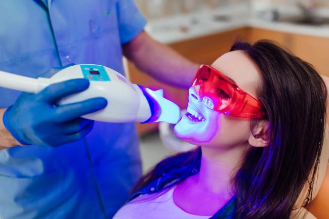 A dental professional performs a teeth whitening procedure on a patient using a blue light device.