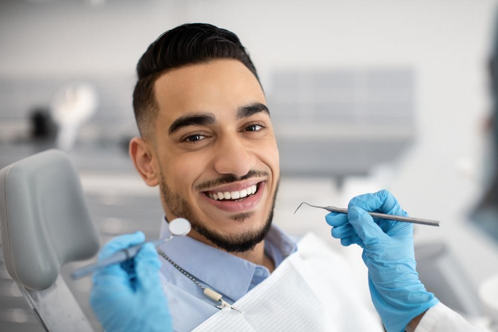 Man in dental chair smiling, with gloved hands holding dental tools.