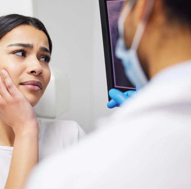 A concerned woman holds her jaw while a masked dentist points to her X-ray on a screen.