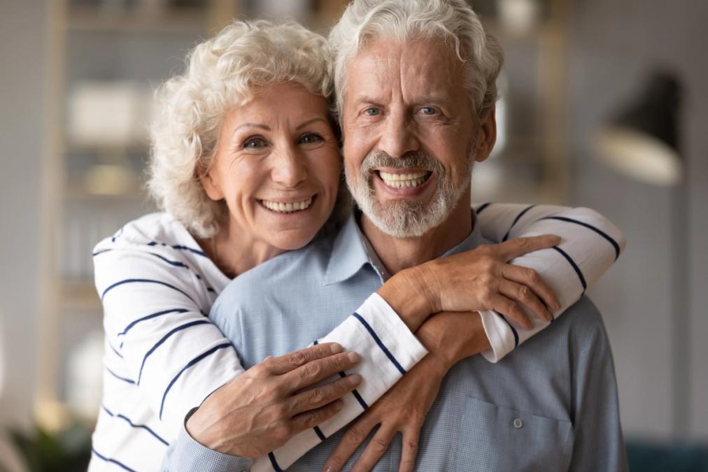 A smiling older woman hugs an older man from behind.