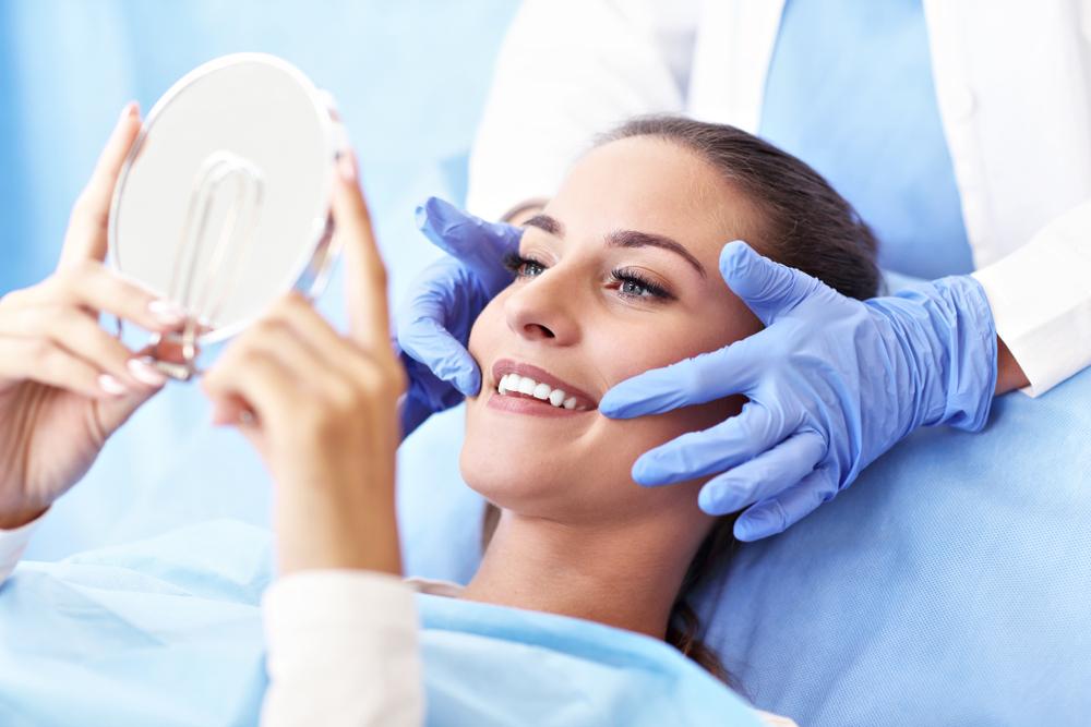 A woman in a dental chair smiles at her bright teeth in a mirror, assisted by a gloved hand.