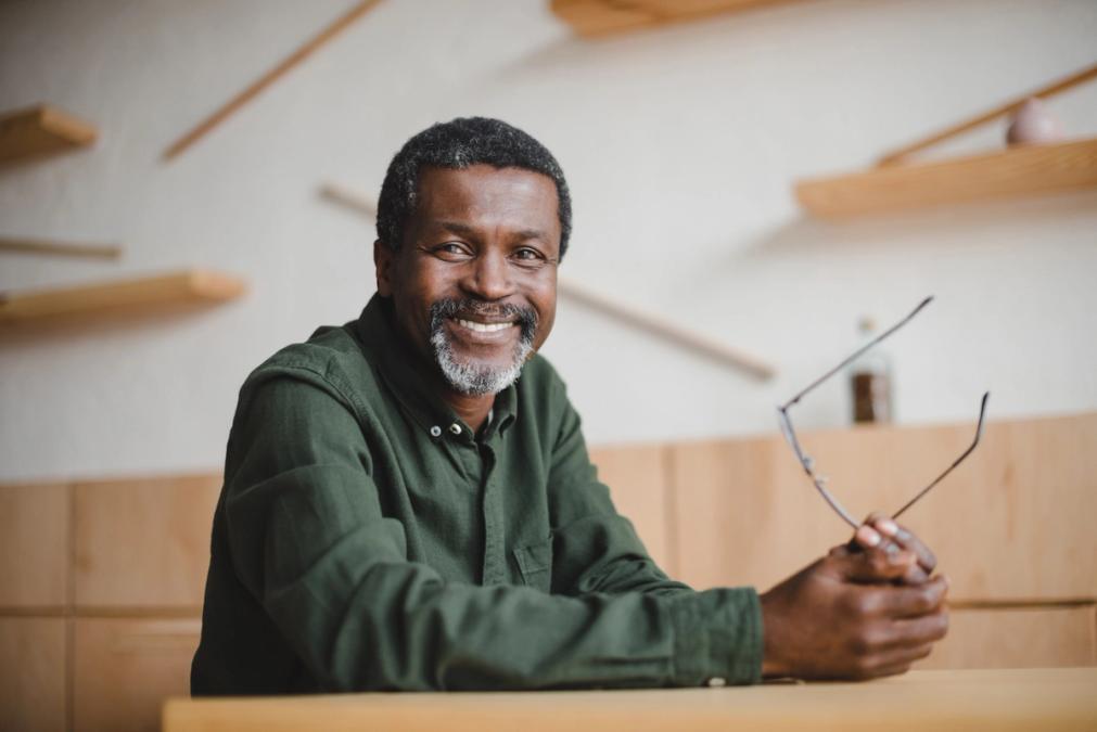 Smiling older Black man with a beard, holding glasses and wearing a green shirt.