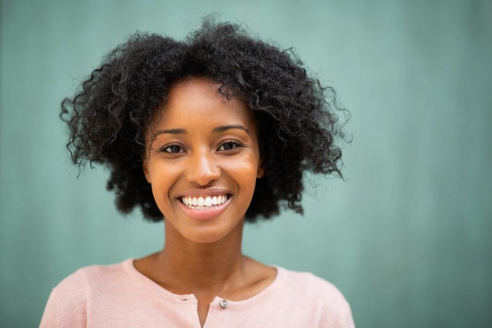 A smiling Black woman with curly hair wearing a pink top against a teal background.
