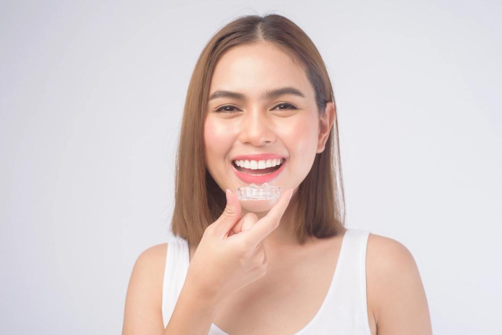 A smiling woman holding a clear dental aligner near her mouth.