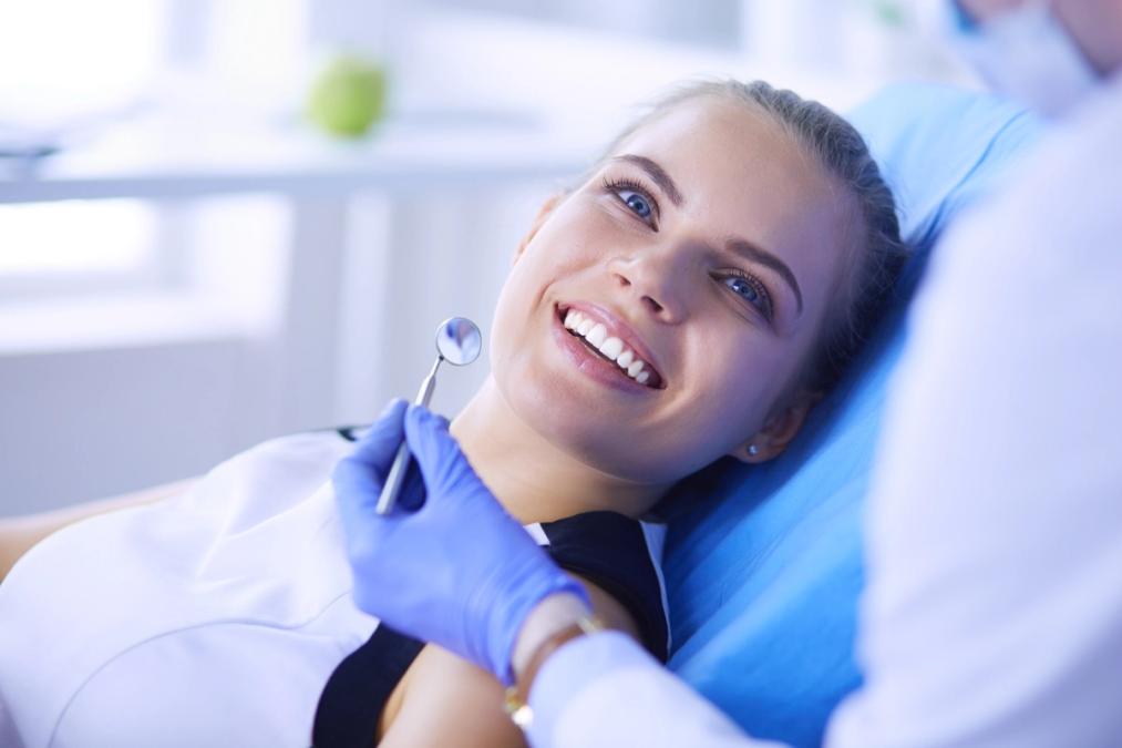 A young woman smiles brightly in a dental chair while a gloved hand holds a dental mirror near her mouth.
