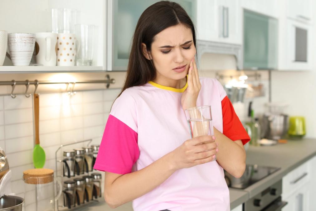 A woman in a kitchen holding a glass of water, grimacing and holding her cheek in pain.
