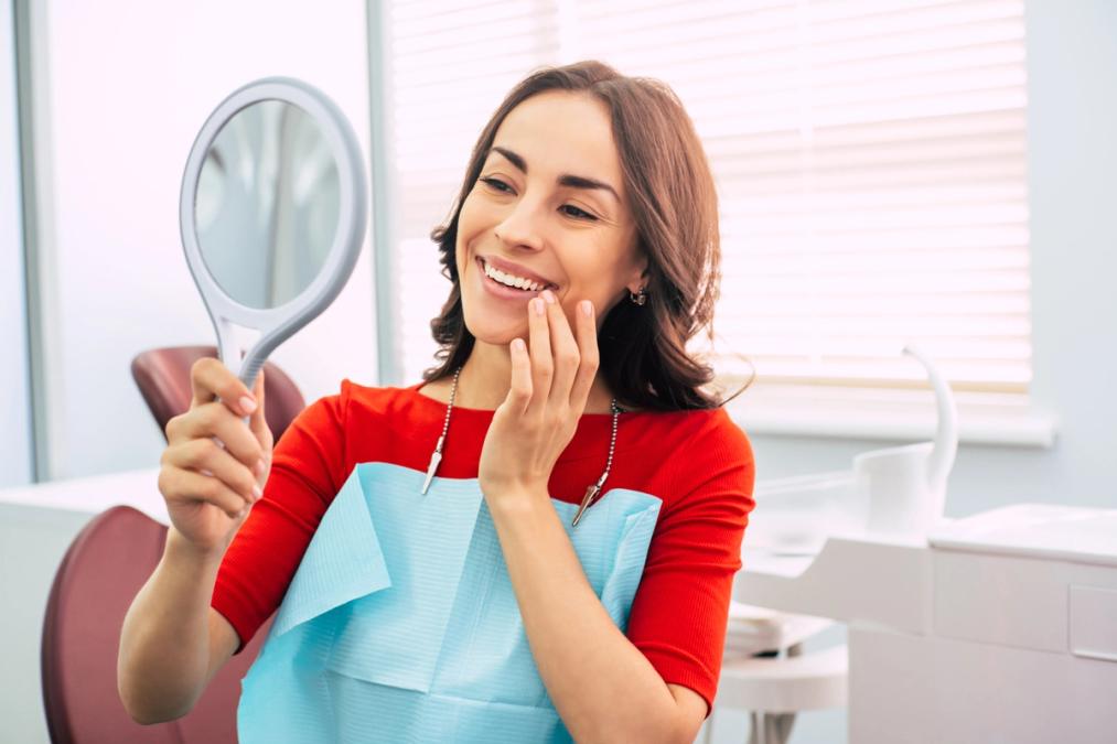 A smiling woman in a dental office looks at her teeth in a mirror.