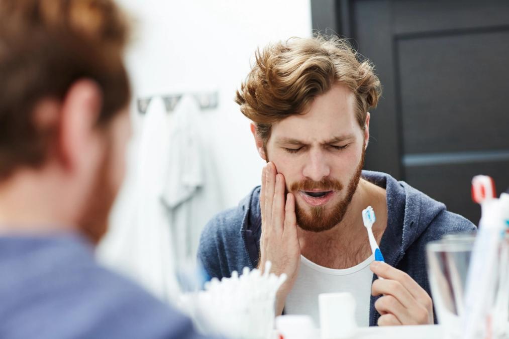 A bearded man looking in a mirror, grimacing in pain and holding his cheek while holding a toothbrush.